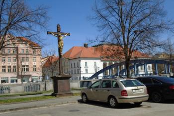 kříž na Zátkově nábřeží (u řeky Malše) od jihu, vpozadí Zlatý most a budova Jihočeského divadla (© Marcela Váňová  04/2021)
